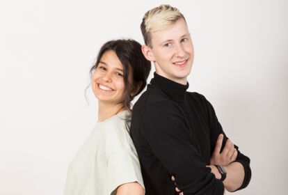 Eine junge Frau und ein junger Mann stehen Rücken an Rücken und lächeln in die Kamera.