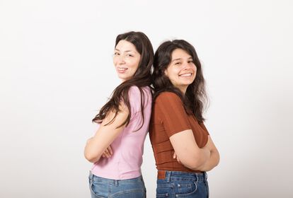Zwei junge Frauen stehen Rücken an Rücken und lächeln in die Kamera.